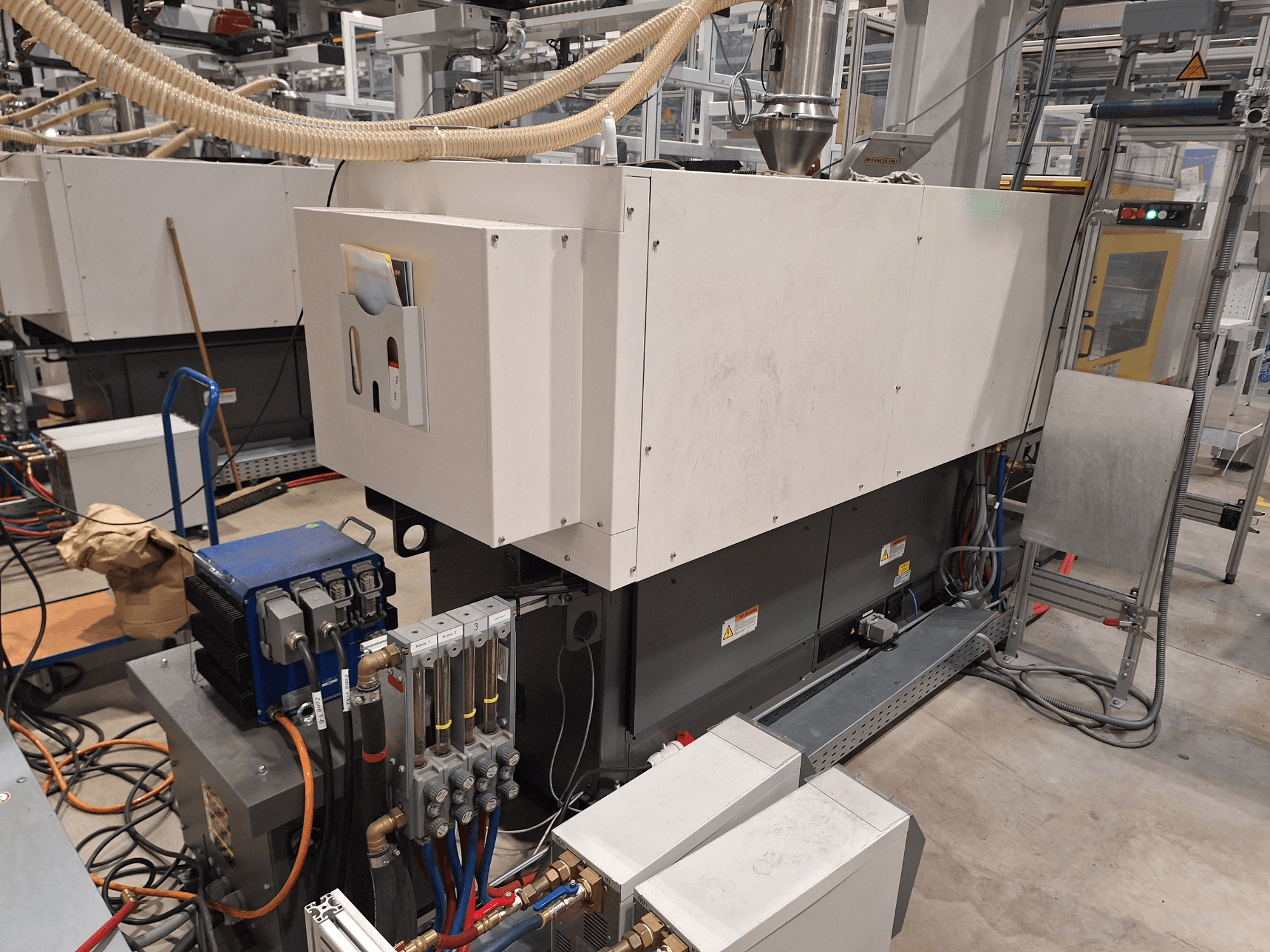 Промислове обладнання в заводських умовах; включає велику машину з панеллю керування, шлангами та електричними з'єднаннями, вид збоку.