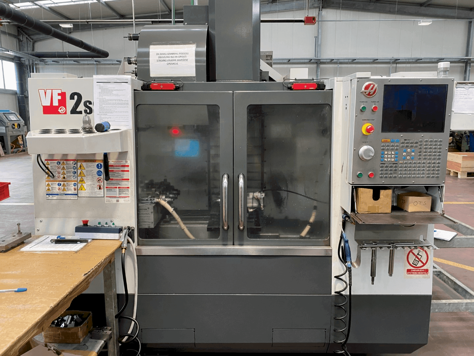 Обробний центр Haas VF-2SS спереду, з панеллю керування, шлангом для охолоджувальної рідини та етикетками безпеки збоку.